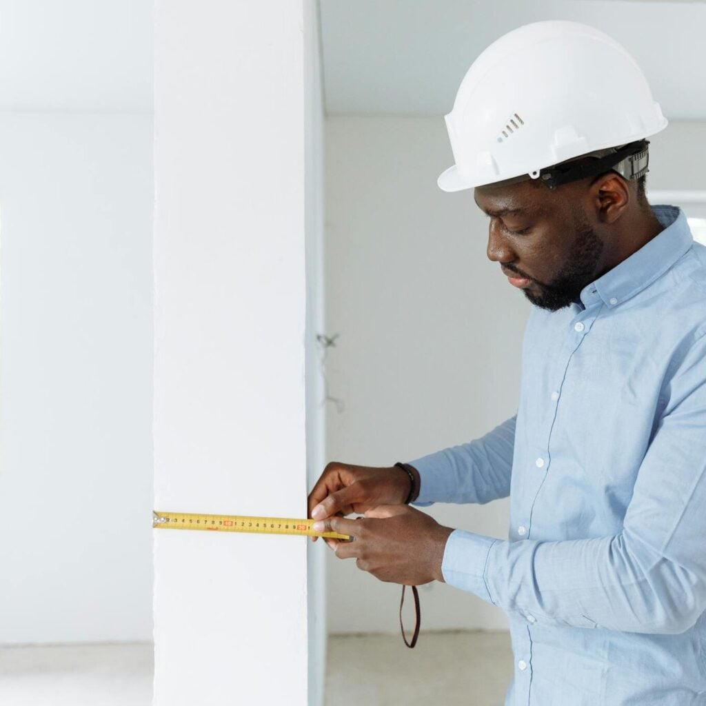 Construction worker measuring a wall indoors, wearing a white hard hat and blue shirt.